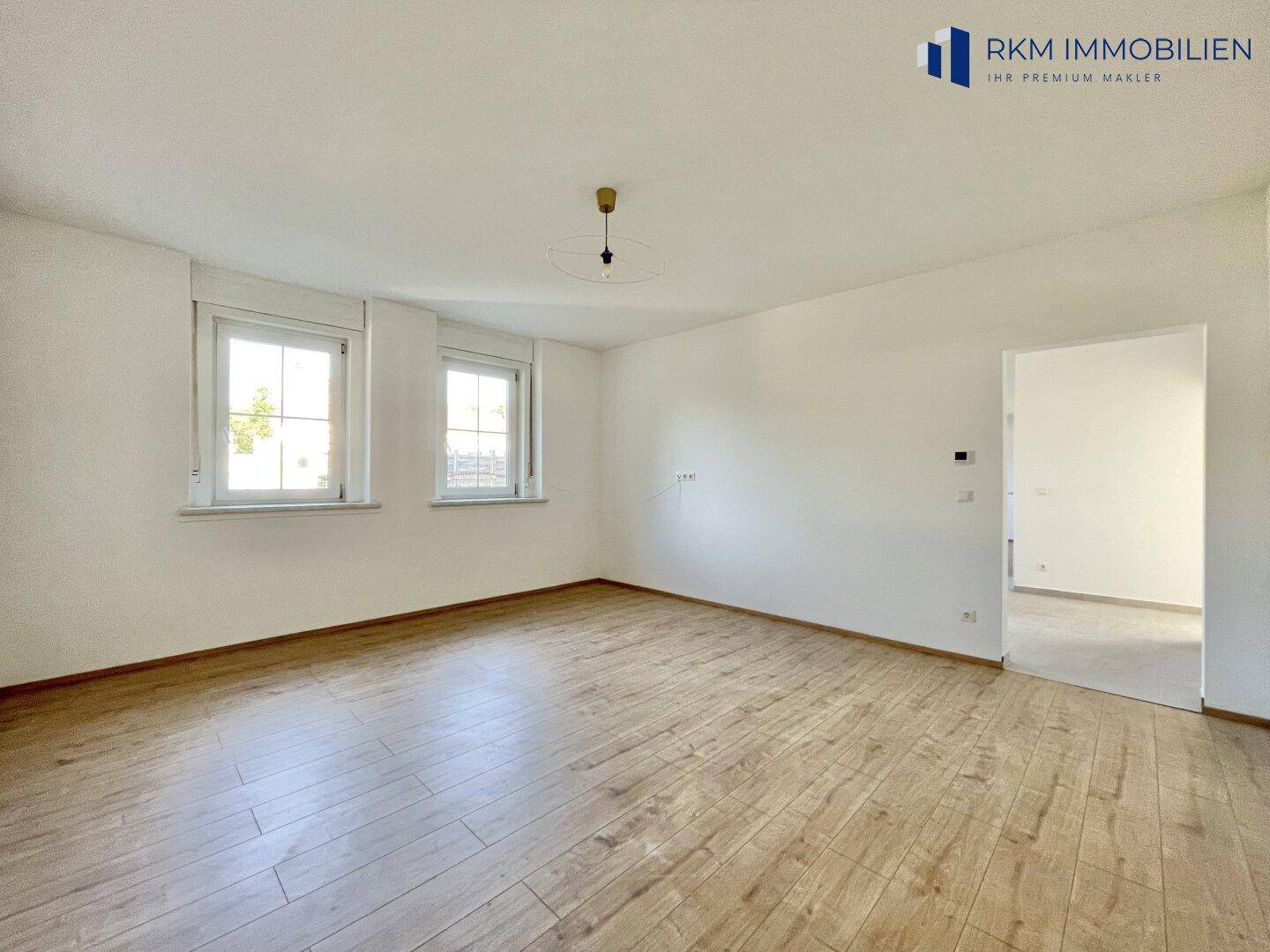 Heller Raum mit Holzboden, zwei Fenstern und einer modernen Deckenleuchte.