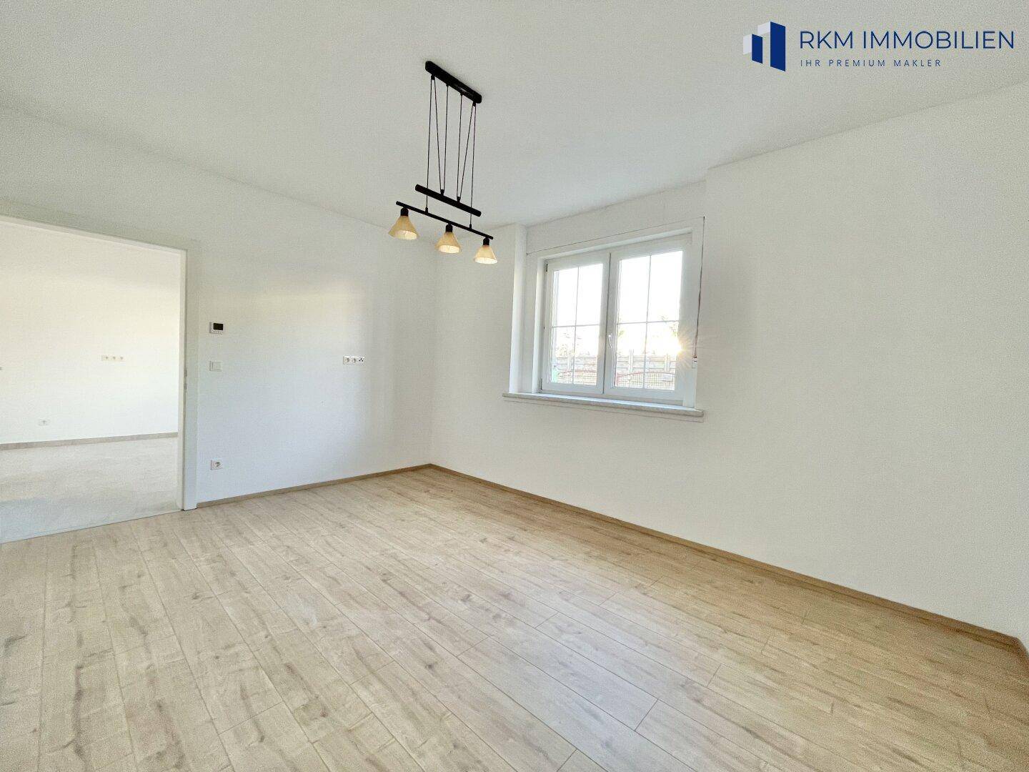 Lichtdurchfluteter Raum mit Holzboden und moderner Hängelampe über dem Fensterbereich.