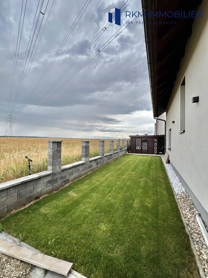 Langer, schmaler Gartenbereich mit Rasen und einer Steinmauer, angrenzend an ein Feld.