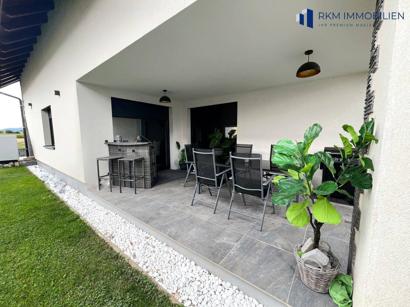 Geräumige überdachte Terrasse mit modernen Gartenmöbeln und einem großen Pflanzgefäß.