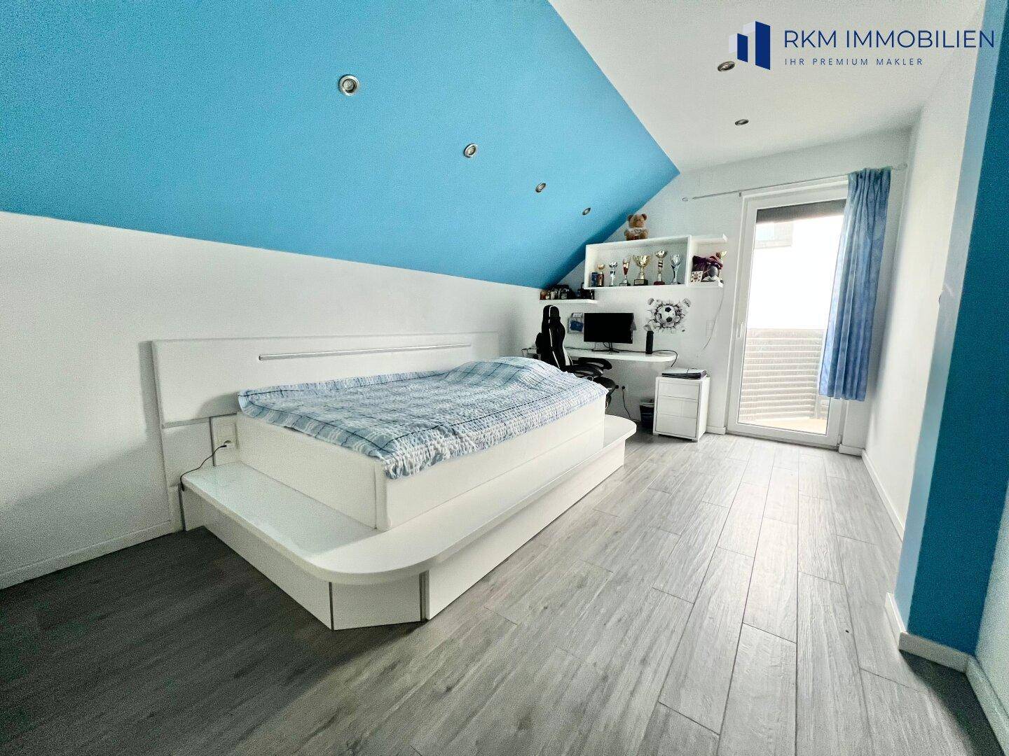 Jugendzimmer mit blau gestrichener Dachschräge, großem Bett und Schreibtischbereich.