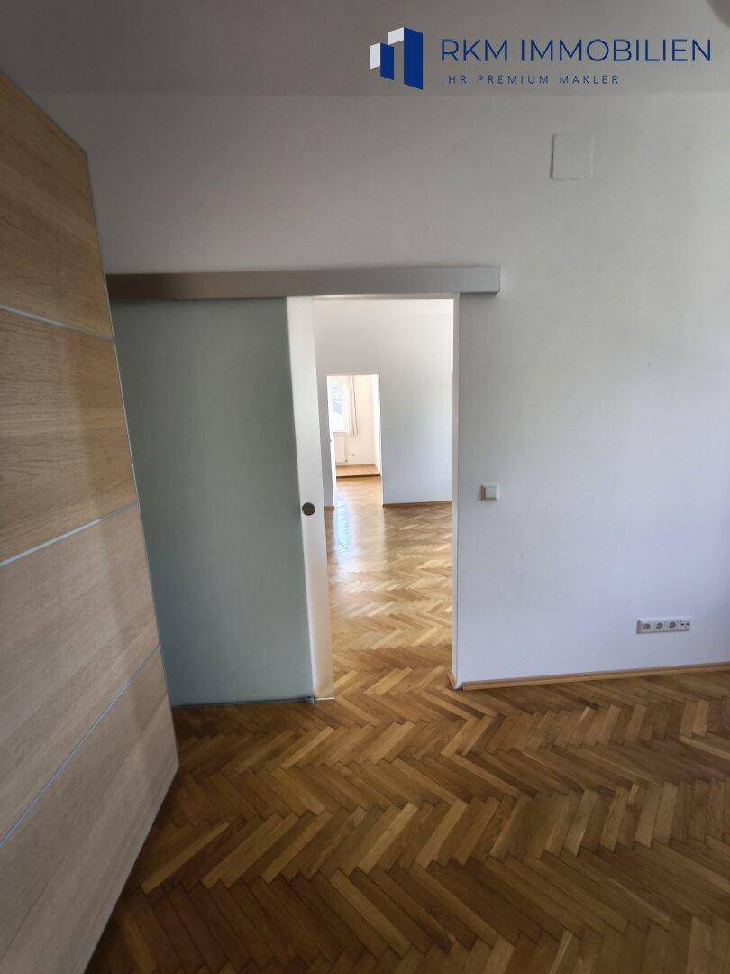 Heller Raum mit Fischgrätparkett und einer Glasschiebetür, die den Übergang zu anderen Bereichen schafft.
