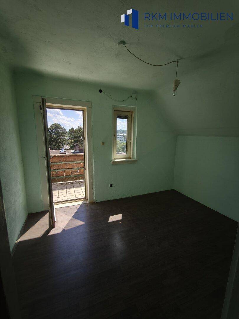 Einfacher Raum mit schräger Decke, Fenster und Zugang zu einem kleinen Balkon.