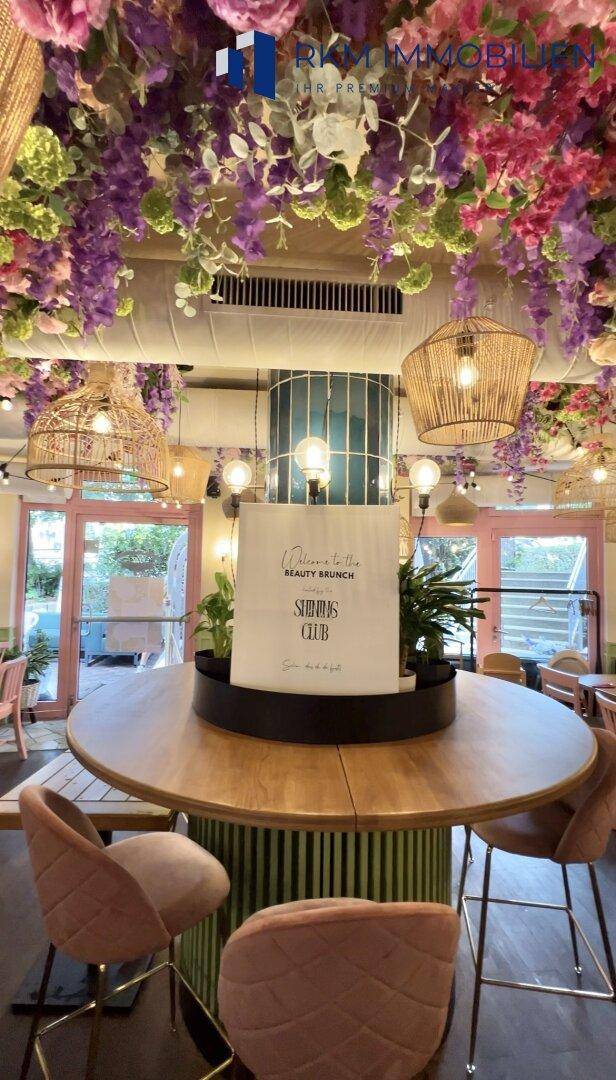 Runder Tisch mit bequemen Stühlen in einem hellen Café mit Blumen an der Decke und Korblampen.