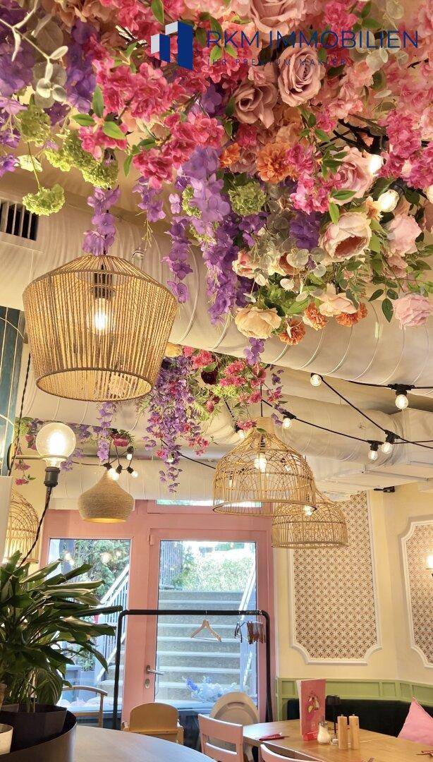 Innenansicht eines Cafés mit üppiger Blumendekoration an der Decke und Korblampen.