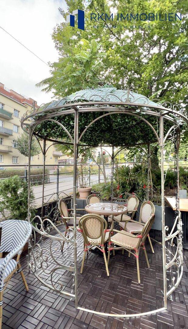 Dekorativer Pavillon mit Sitzgelegenheiten auf einer Außenterrasse, umgeben von Bäumen.