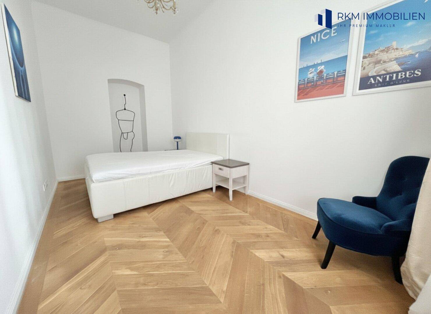 Helles Schlafzimmer mit modernem Bett, blauem Sessel und elegantem Fischgrätparkettboden.