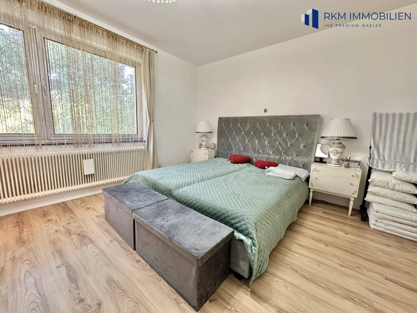Helles Schlafzimmer mit Doppelbett, Holzfußboden und großen Fenstern mit Blick ins Grüne.