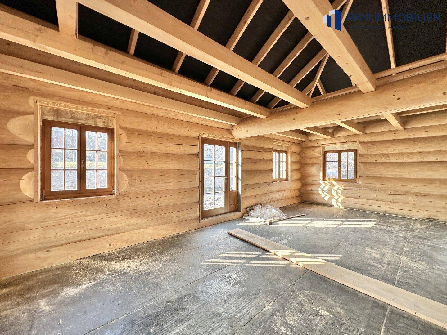 Unfertiger Innenraum eines Blockhauses mit sichtbaren Holzbalken, großen Fenstern und viel Tageslicht.