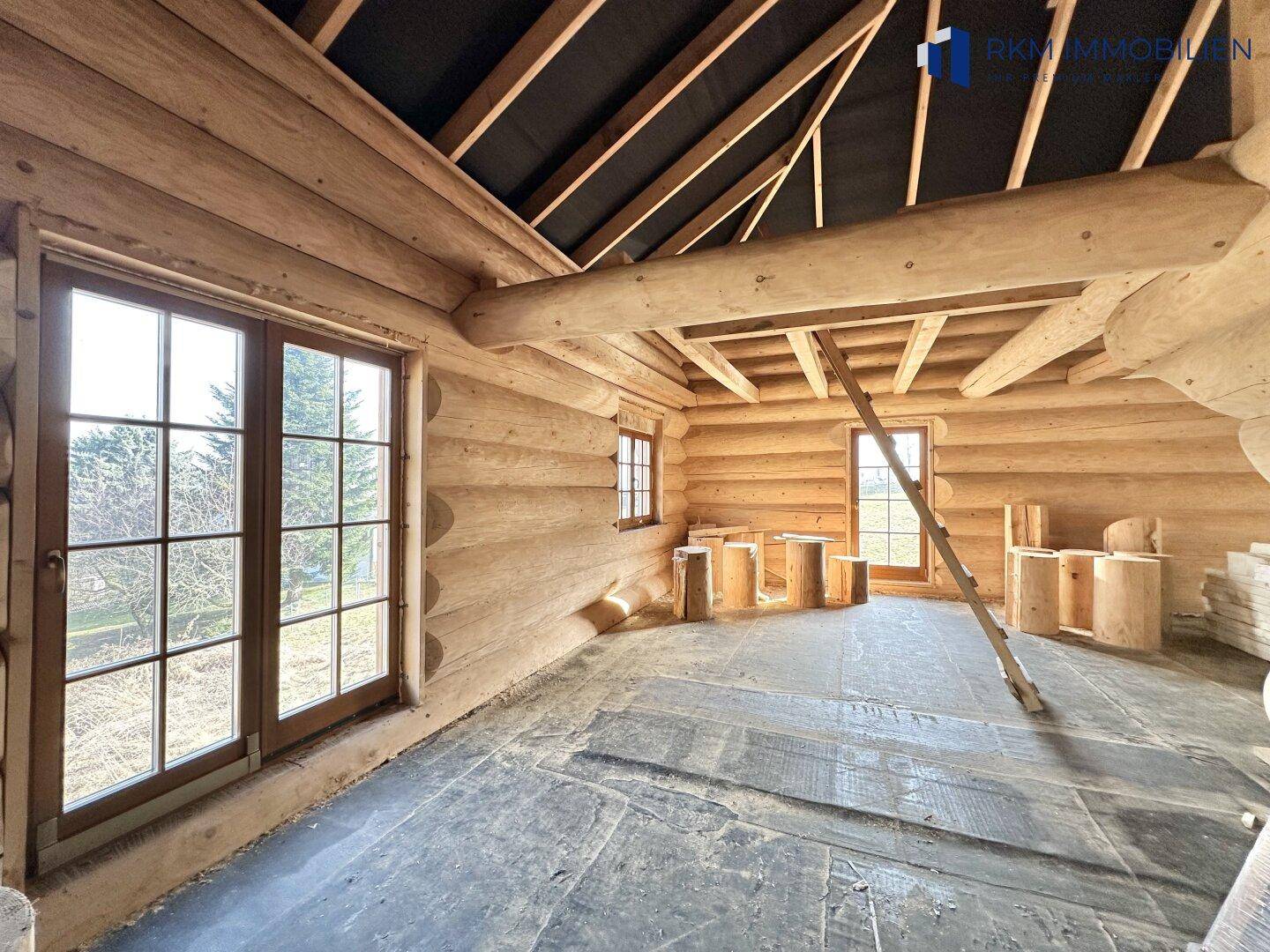 Helle Ecke eines unfertigen Raumes mit großen Sprossenfenstern und charakteristischen Holzstammwänden.