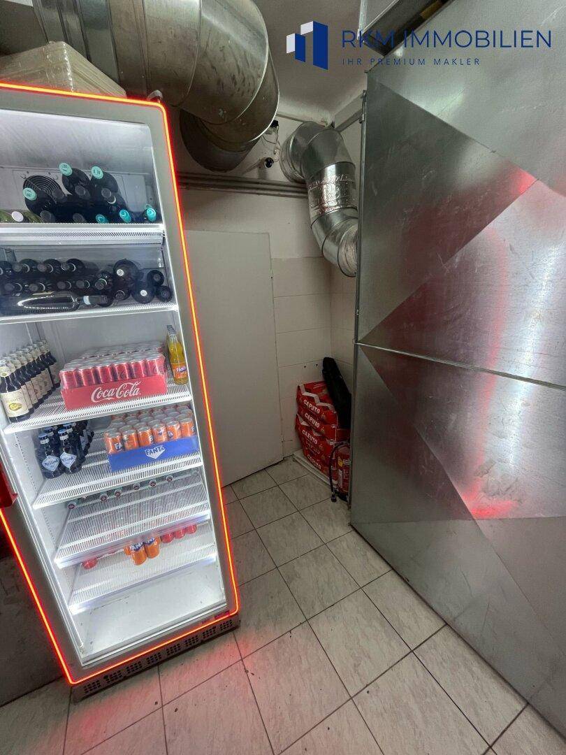 Lagerbereich mit beleuchtetem Getränkekühlschrank und gestapelten Kartons, neben sichtbaren Lüftungsrohren.