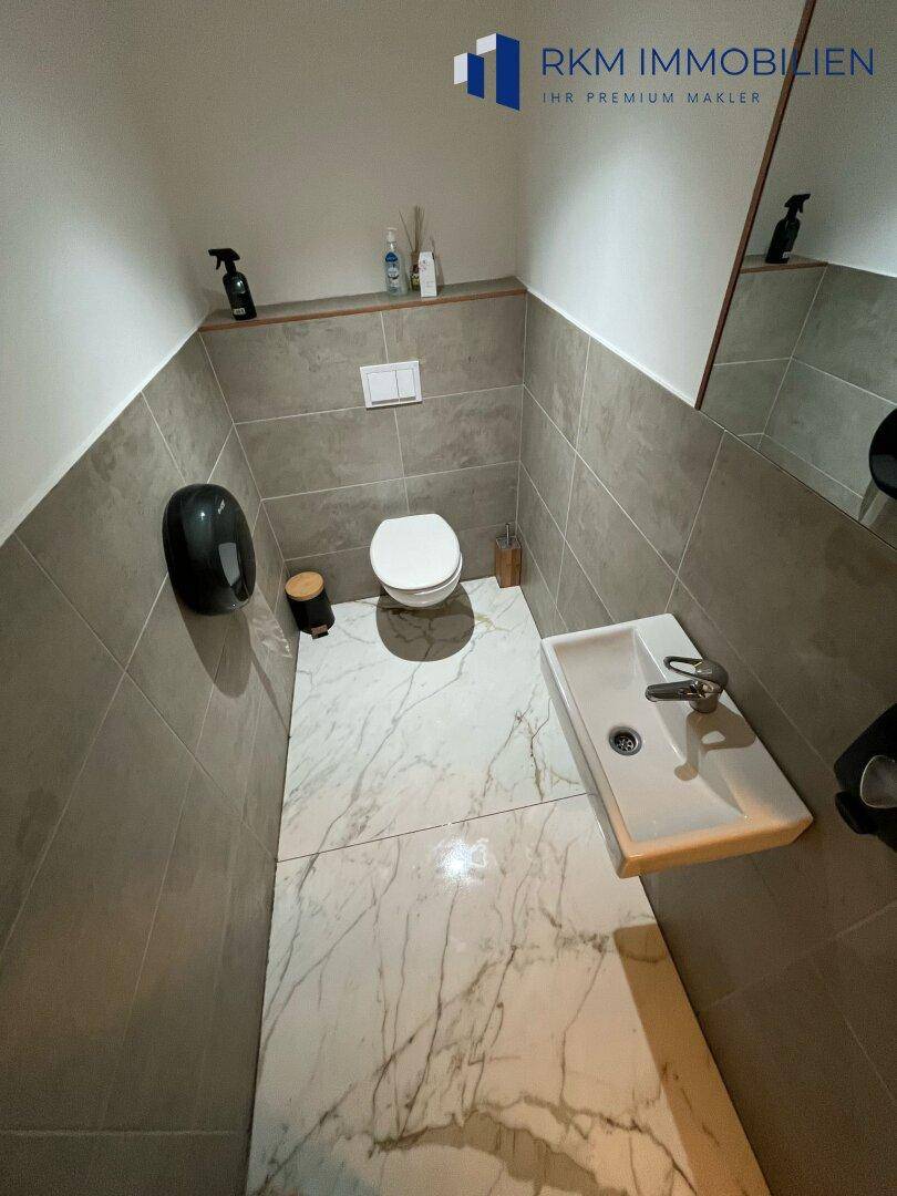 Sauberer Toilettenraum mit Wand-WC, Waschbecken und großem Spiegel, in neutralen Grautönen gehalten, mit Fliesenboden.