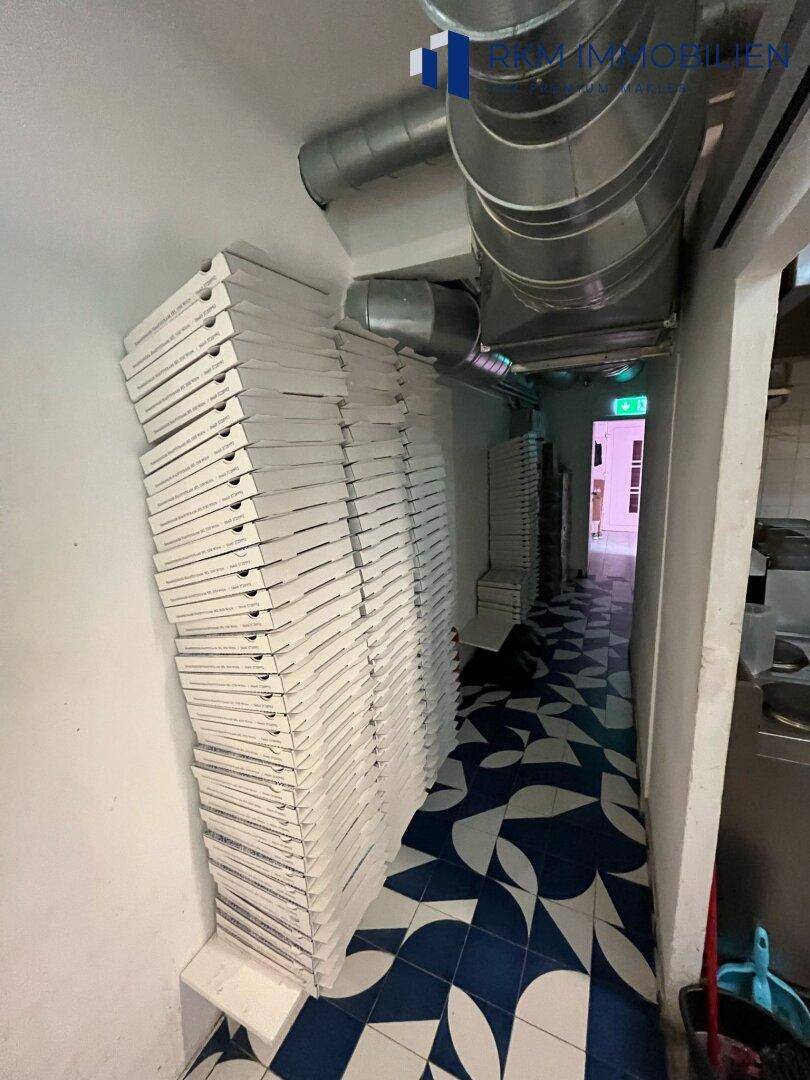 Schmaler Flur mit gestapelten Pizzakartons und sichtbaren Lüftungsrohren, führt zu weiteren Räumen.