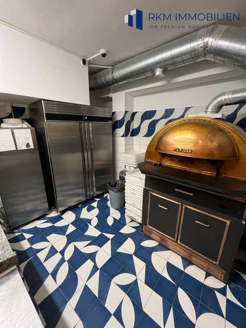 Professionelle Küche mit großem Pizzaofen, zwei Edelstahlkühlschränken und sichtbaren Lüftungsrohren, mit Kamera.