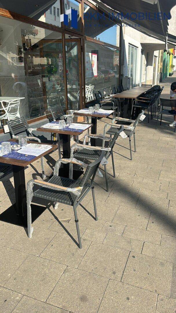 Sonniger Außenbereich mit Tischen und Stühlen auf dem Bürgersteig, vor der Ladenfront, mit natürlichem Licht.