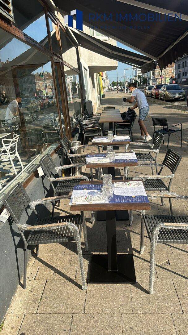 Belebtes Straßencafé mit dunklen Tischen und Stühlen auf dem Bürgersteig, unter einer Markise, mit natürlichem Licht.