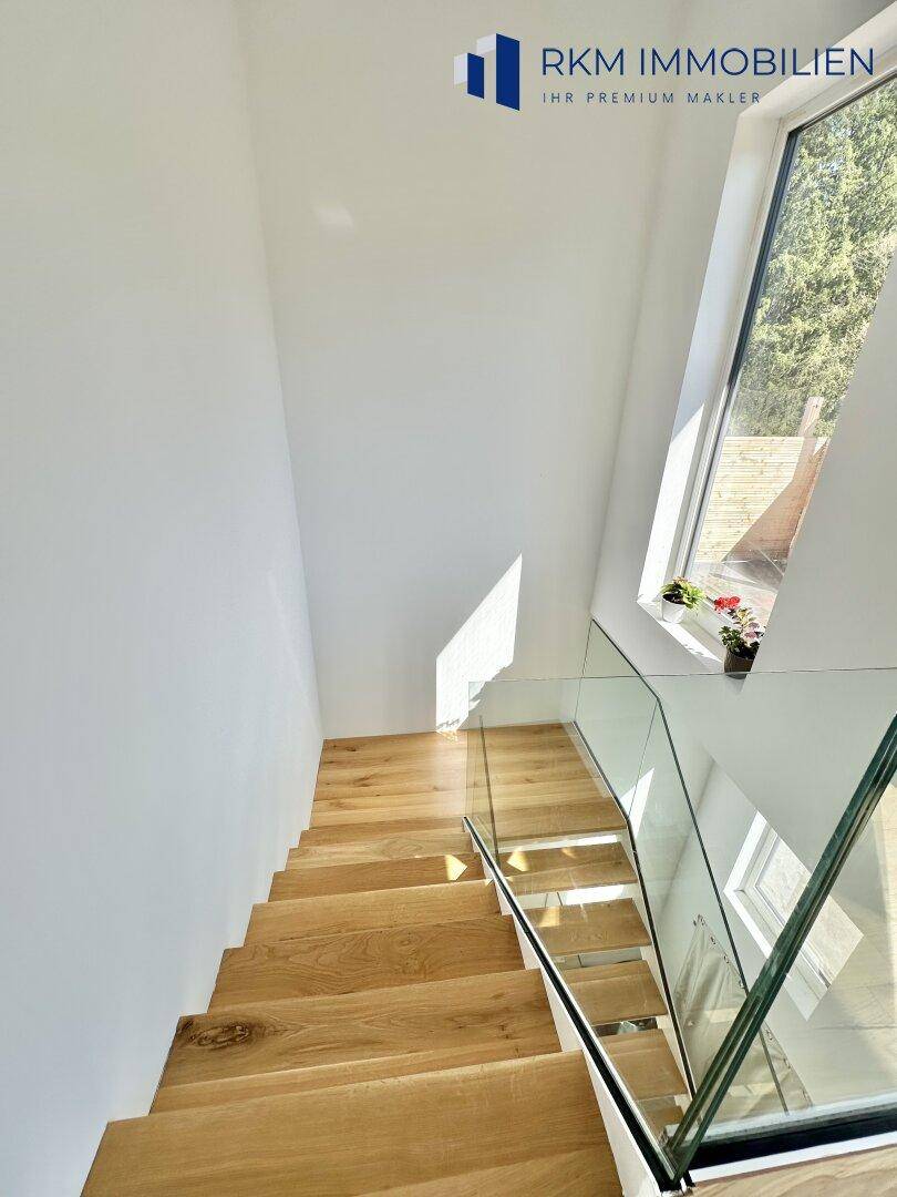 Elegante Holztreppe mit Glasgeländer und großem Fenster, das viel Licht hereinlässt.