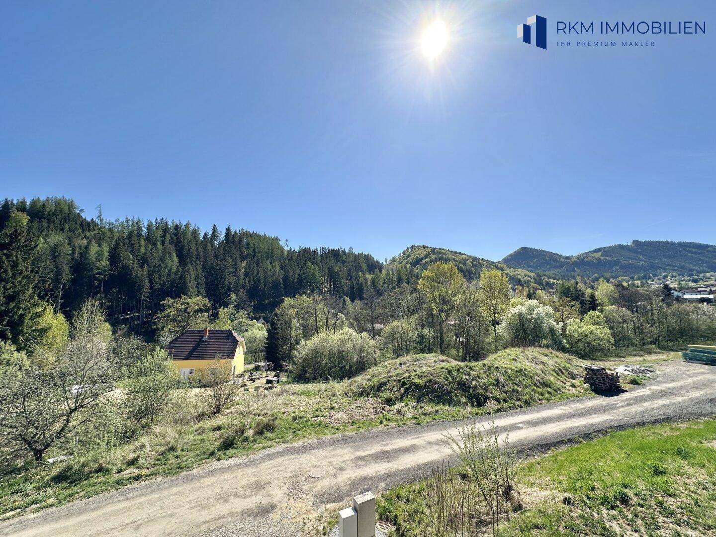 Weitläufiger Blick über eine grüne Hügellandschaft mit Wald und einem sonnigen Himmel.