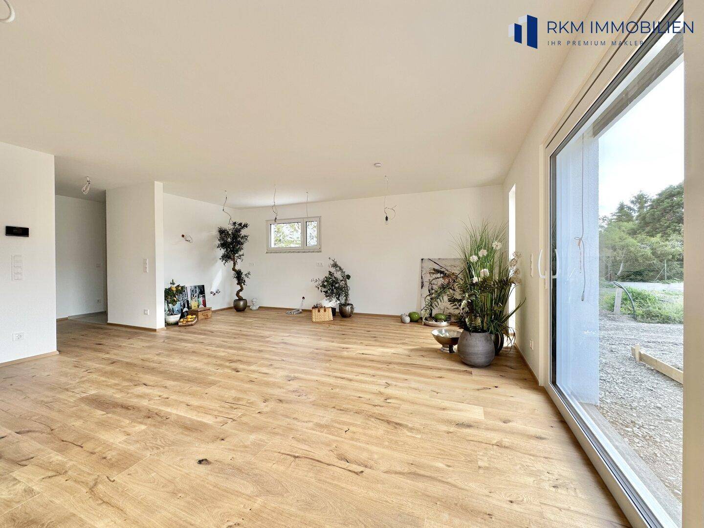 Weitläufiger Wohnbereich mit Holzboden und großen Fenstern, die einen Blick ins Grüne ermöglichen.