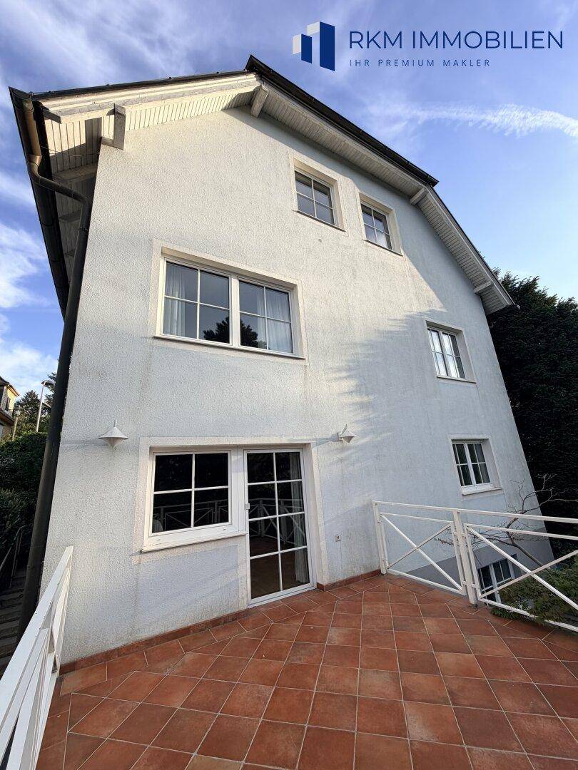 Die helle Hausfassade mit großen Fenstern und einer geräumigen Terrasse mit Terrakottafliesen.