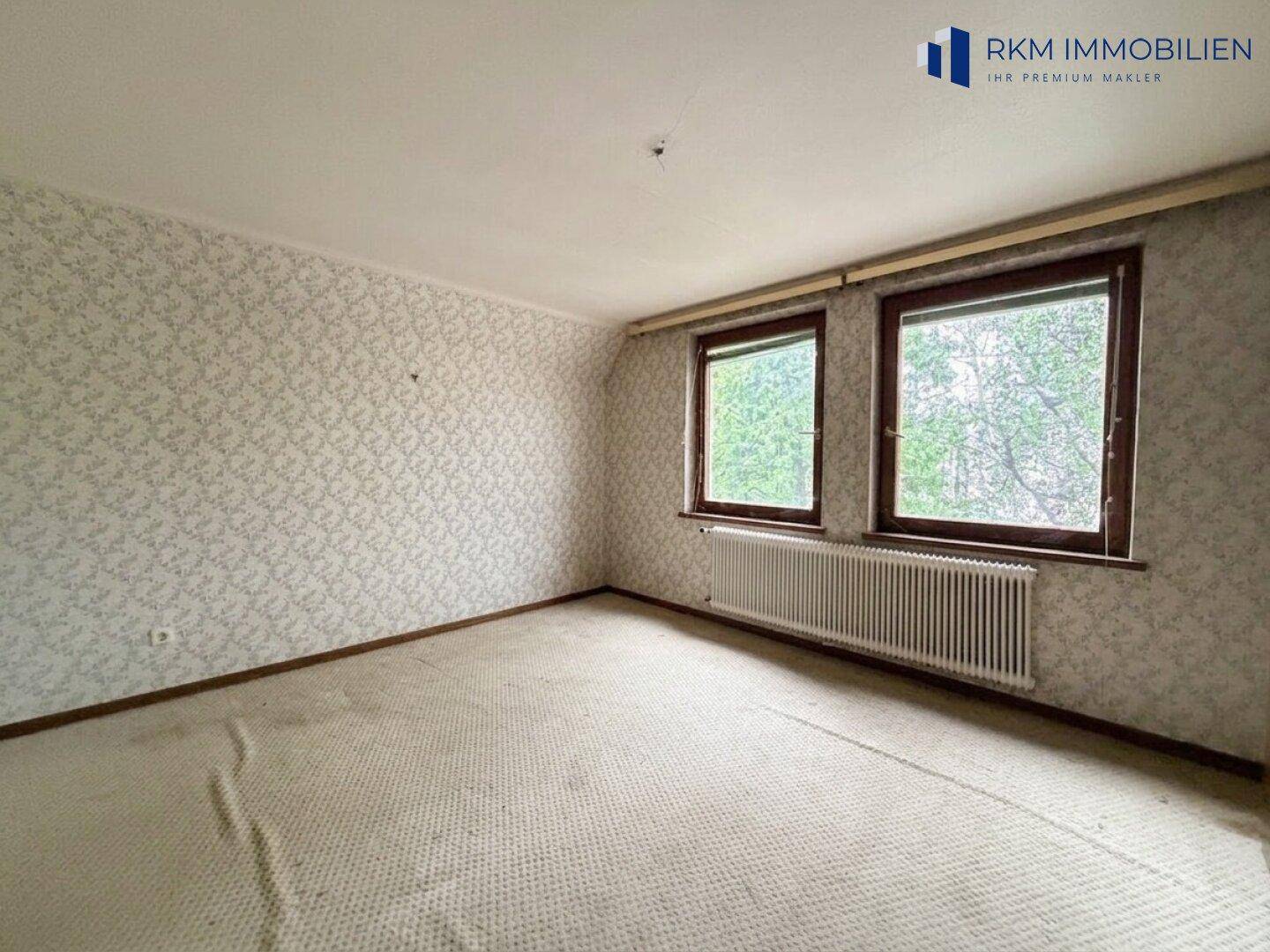 Schlafzimmer mit Teppichboden, zwei Fenstern und gemusterter Tapete.