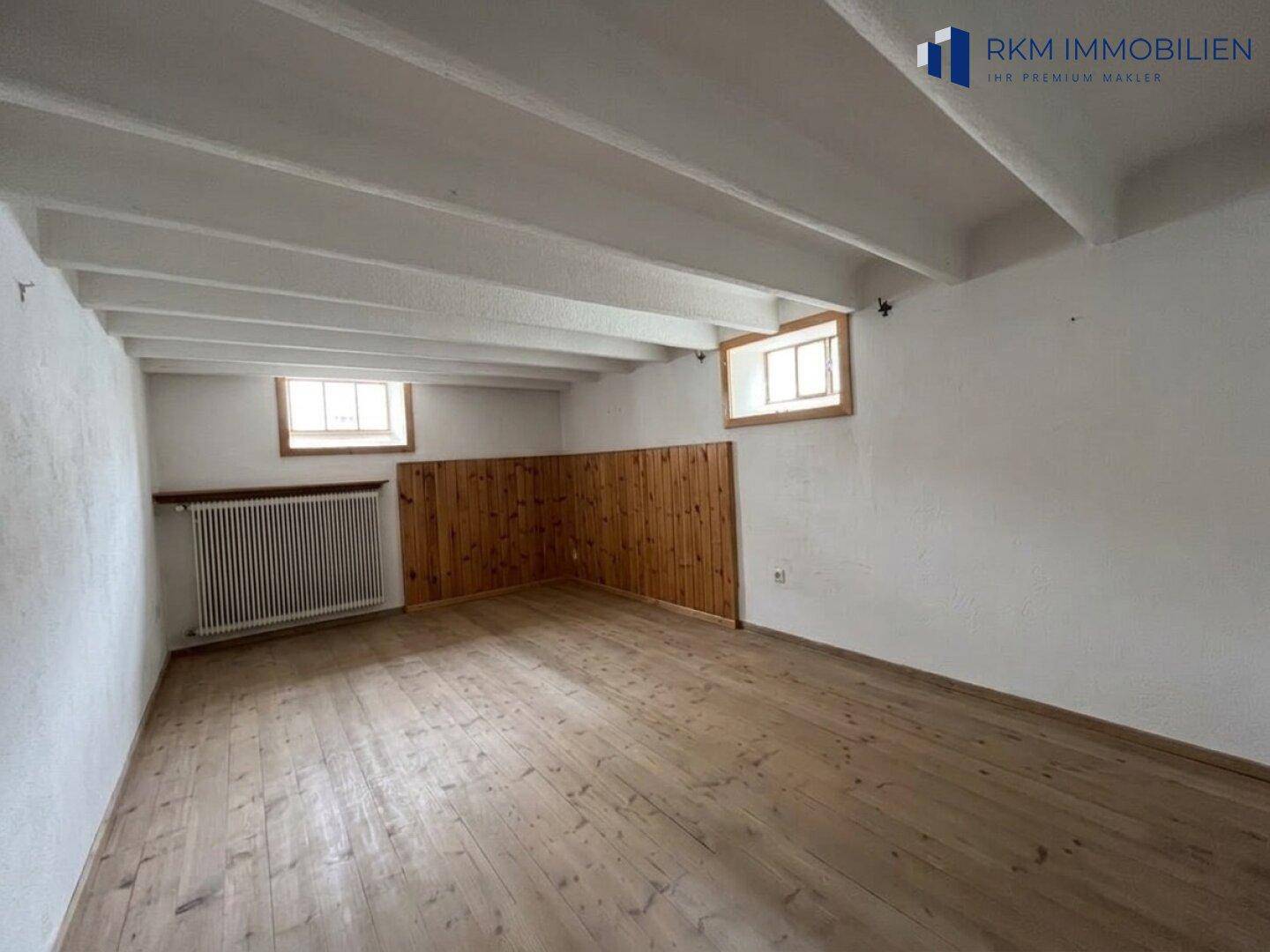 Kellerraum mit Holzbalkendecke, Dielenboden und kleinen Fenstern.