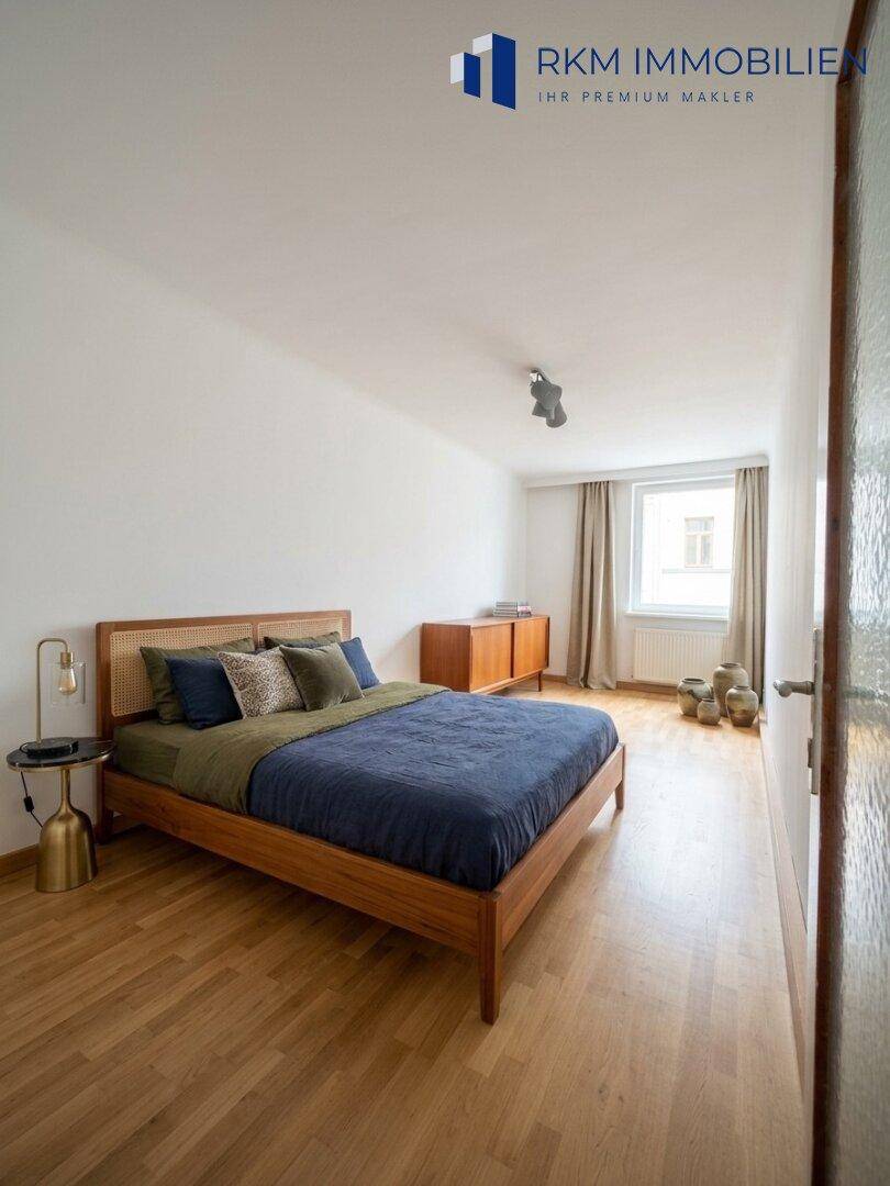 Schlafzimmer mit Holzbett, blauer Bettwäsche und Parkettboden, hell durch großes Fenster.