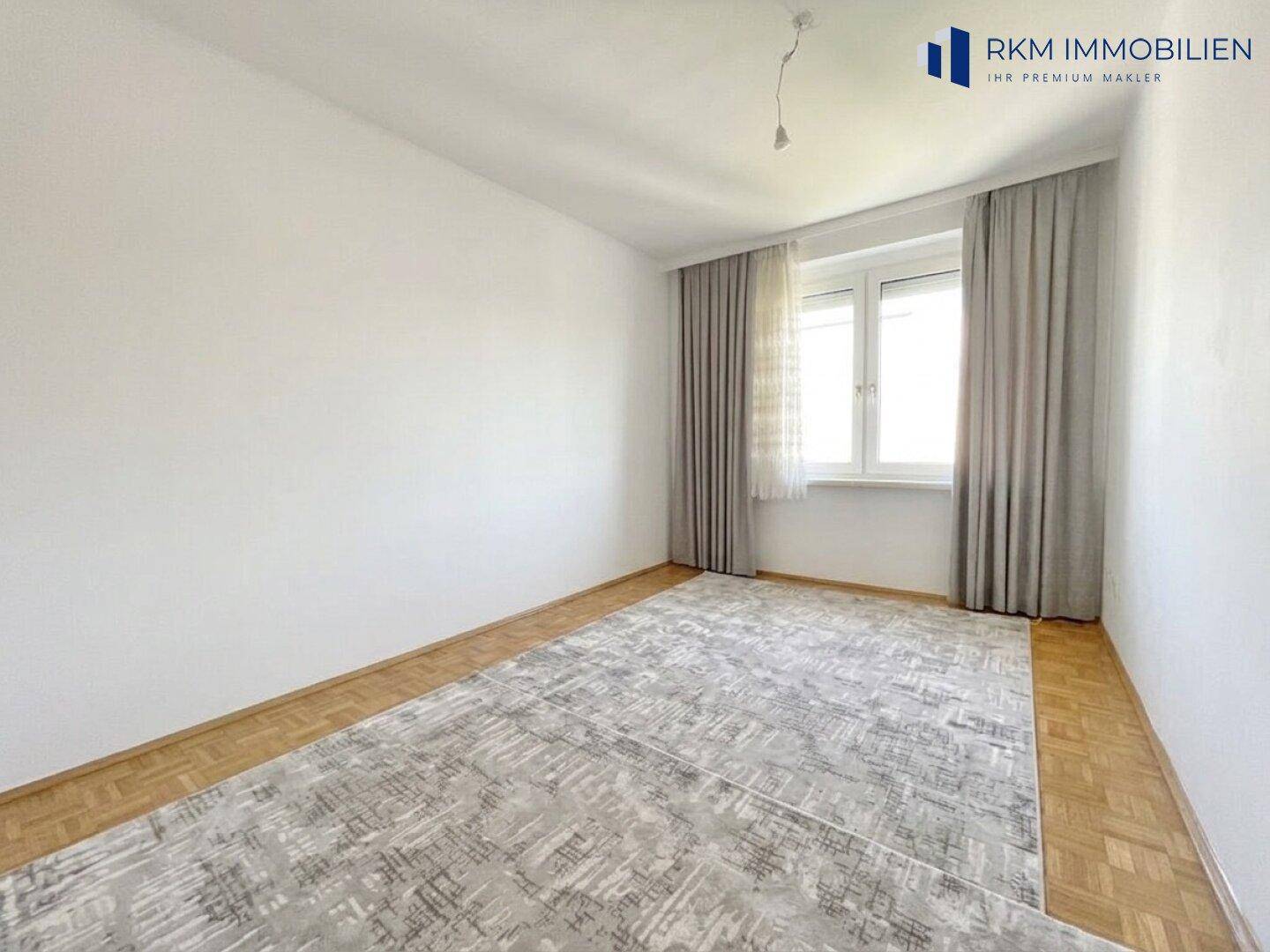 Schlichtes Schlafzimmer mit hellem Holzboden, großem Teppich und Fenster mit Vorhängen.
