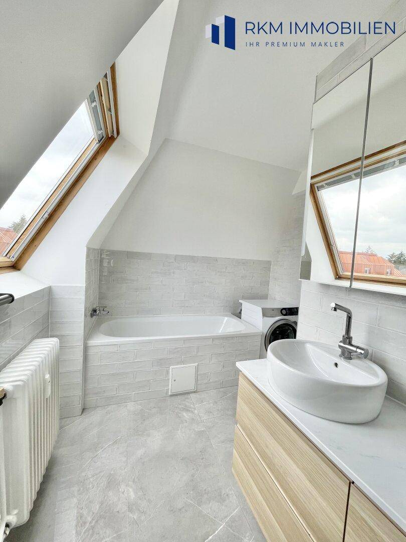 Modernes Badezimmer mit Badewanne, Waschbecken und grauen Fliesen, unter einem Dachfenster gelegen.