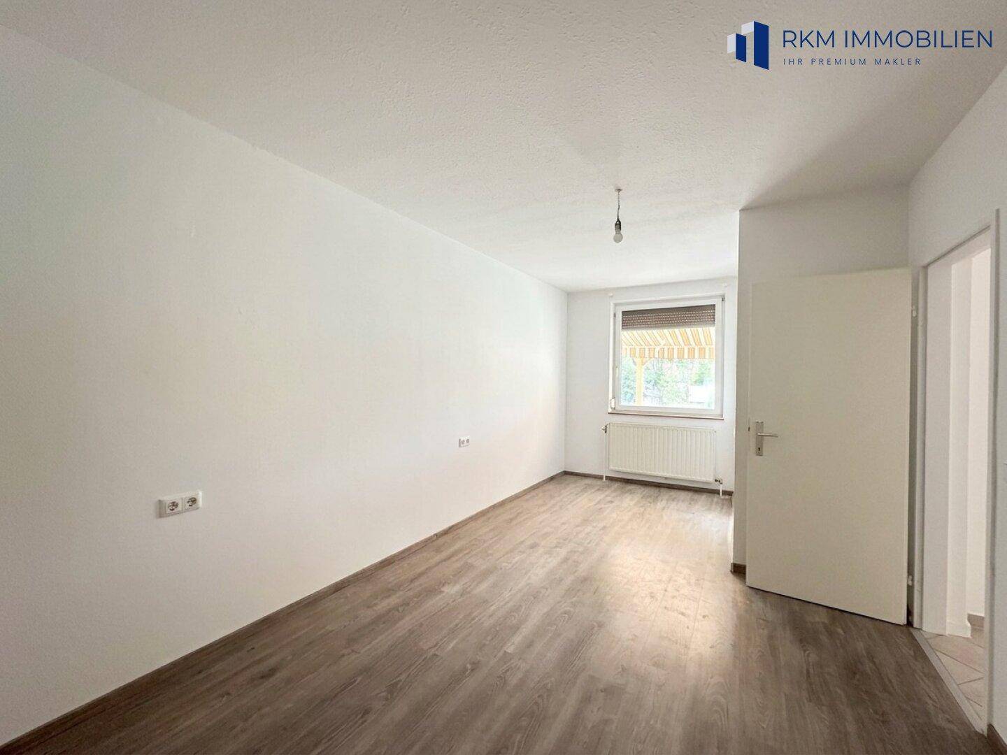 Helles Zimmer mit Laminatboden und einem Fenster mit Rollladen.