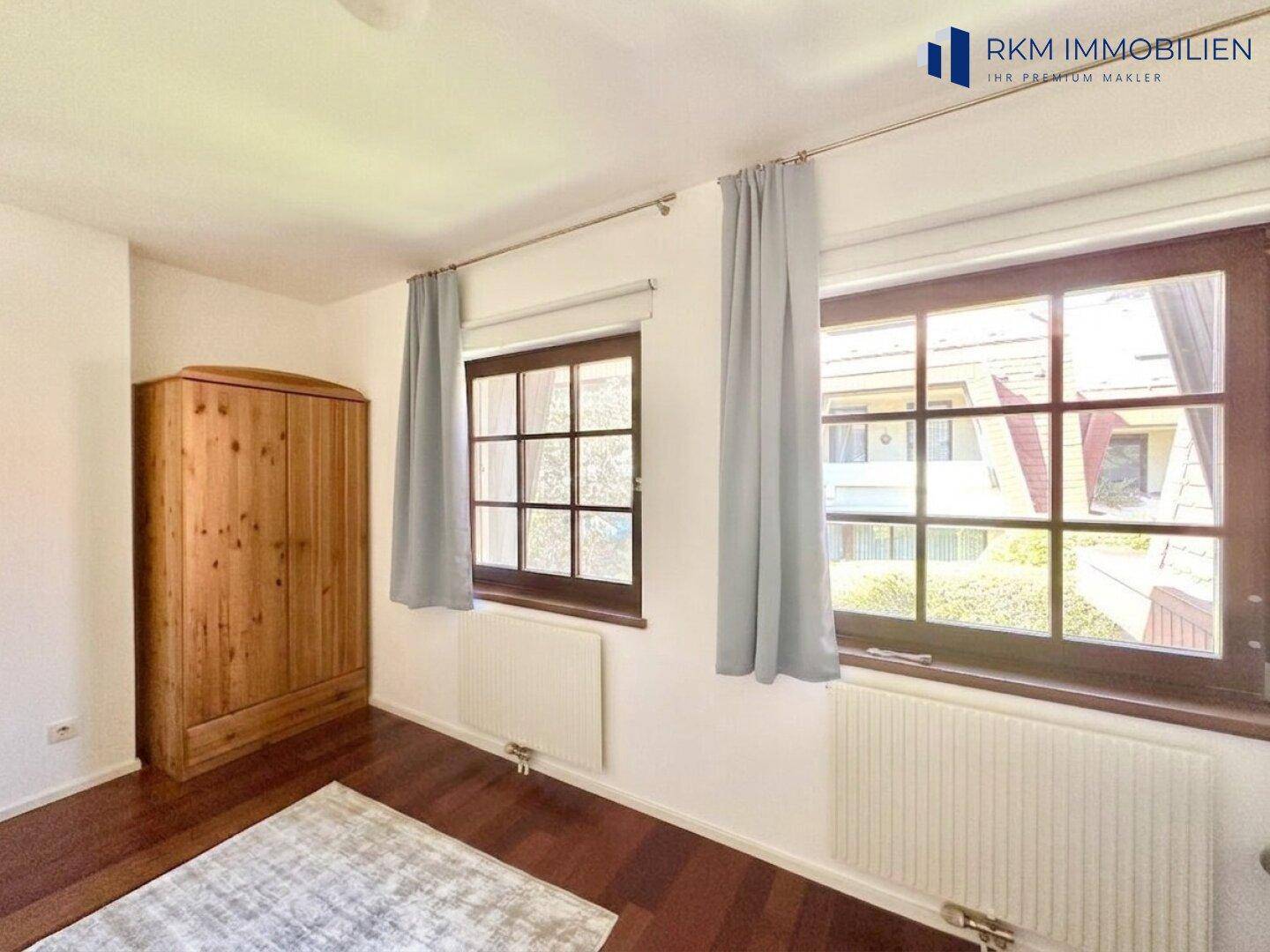 Schlafzimmer mit dunklem Holzboden, großem Holzschrank und zwei Fenstern.