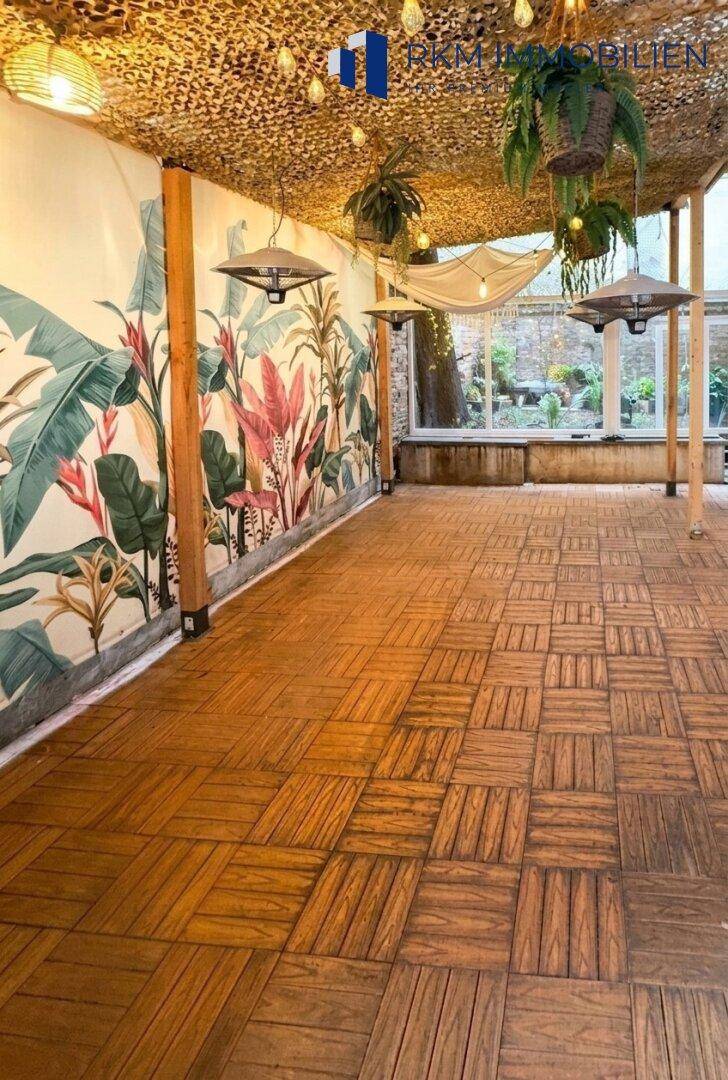 Innenraum mit Holzbodenfliesen, tropischer Wandtapete und hängenden Pflanzen, ideal für Veranstaltungen.