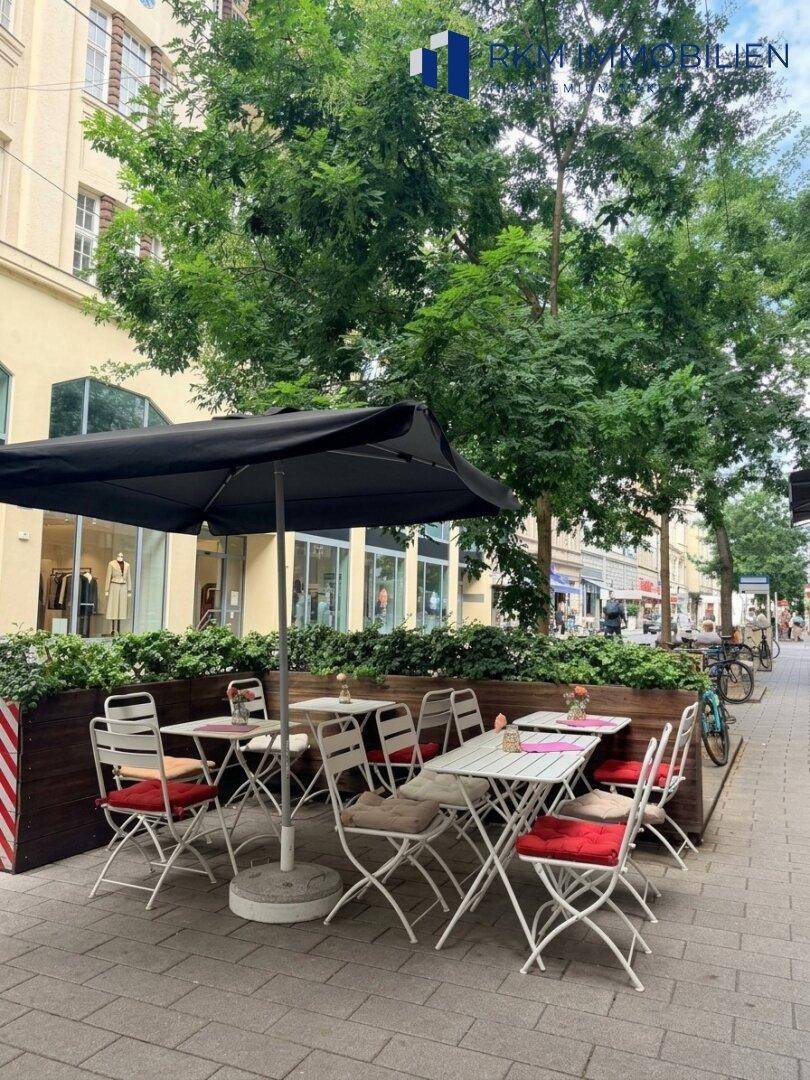 Gemütlicher Außenbereich mit Tischen, Stühlen und Sonnenschirm auf dem Bürgersteig unter Bäumen.