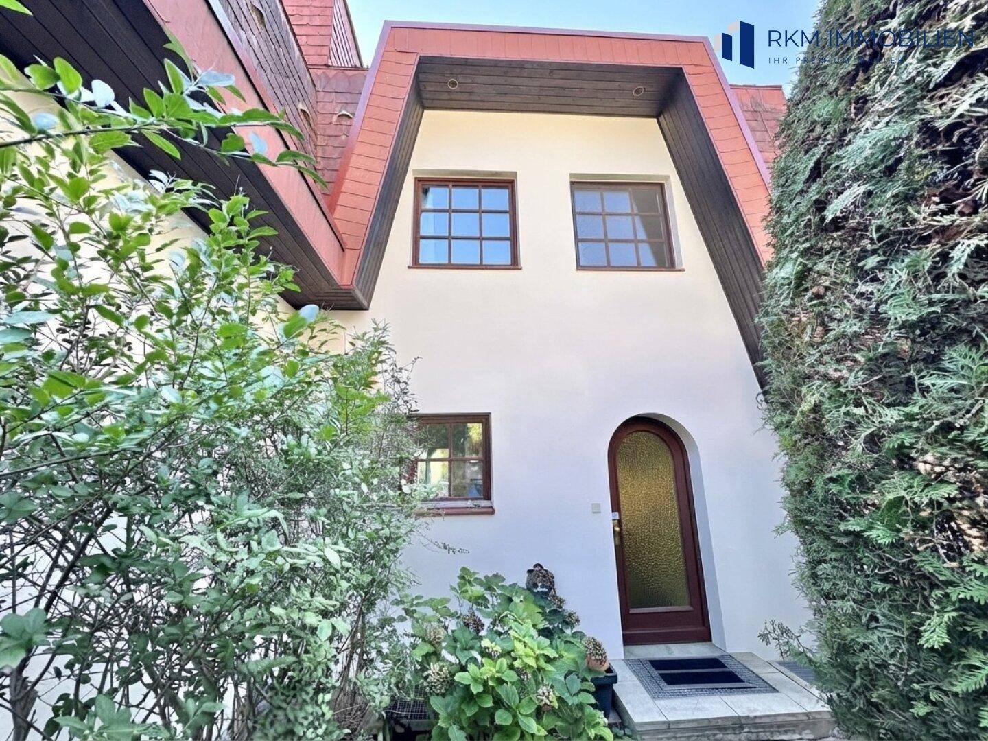 Vorderansicht eines gepflegten Hauses mit weißer Fassade, braunem Dach und üppiger Bepflanzung.