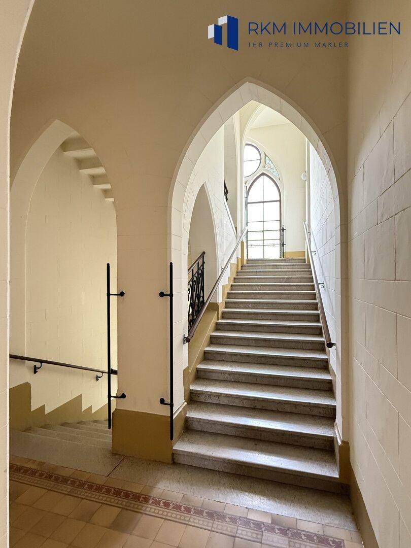 Historisches Treppenhaus mit hohen Spitzbogenfenstern, eleganten Bögen und Steinfliesenboden.