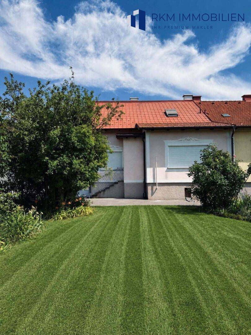 Seitenansicht des Hauses mit frisch gemähtem Rasen und üppiger Bepflanzung unter blauem Himmel.