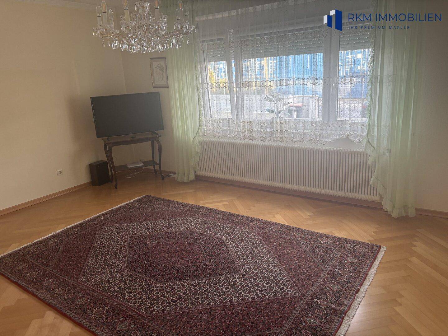Geräumiges Wohnzimmer mit Fischgrätparkett, großem Teppich und breiten Fenstern für viel Tageslicht.