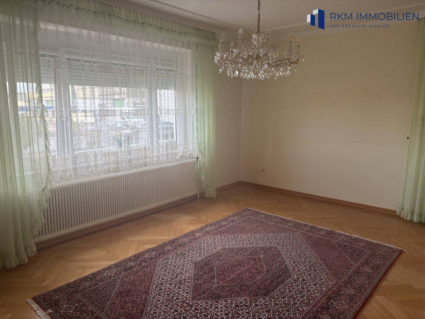 Helles Wohnzimmer mit Parkettboden, einem großen Fenster und einem eleganten Kronleuchter an der Decke.