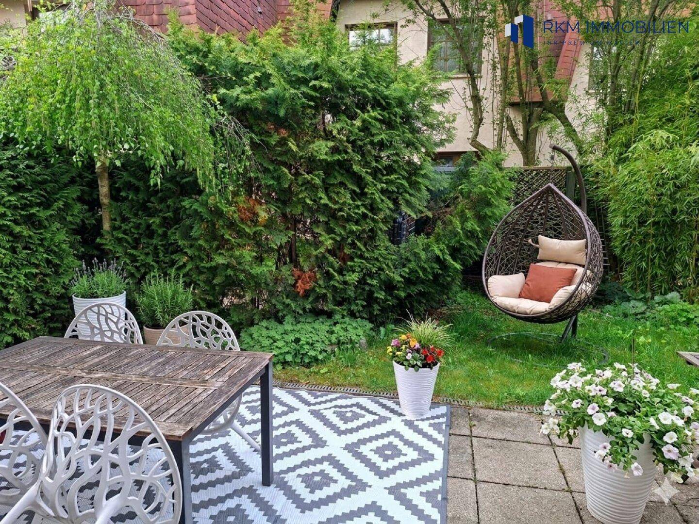 Gemütliche Terrasse mit Sitzgelegenheiten und Hängesessel im grünen Garten, umgeben von üppiger Vegetation.