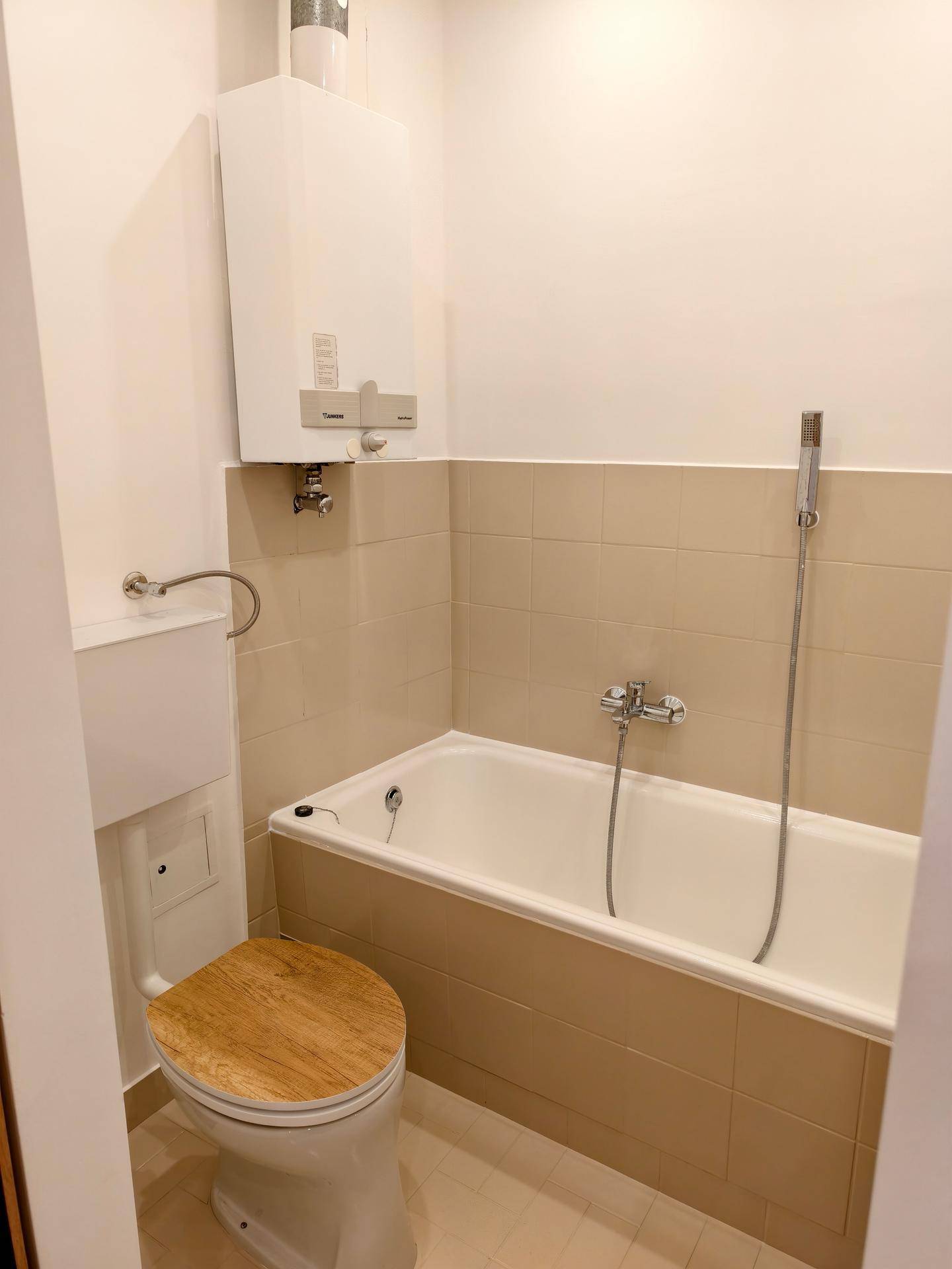 Badezimmer mit Badewanne, WC mit Holzdeckel und beigefarbenen Fliesen an den Wänden.