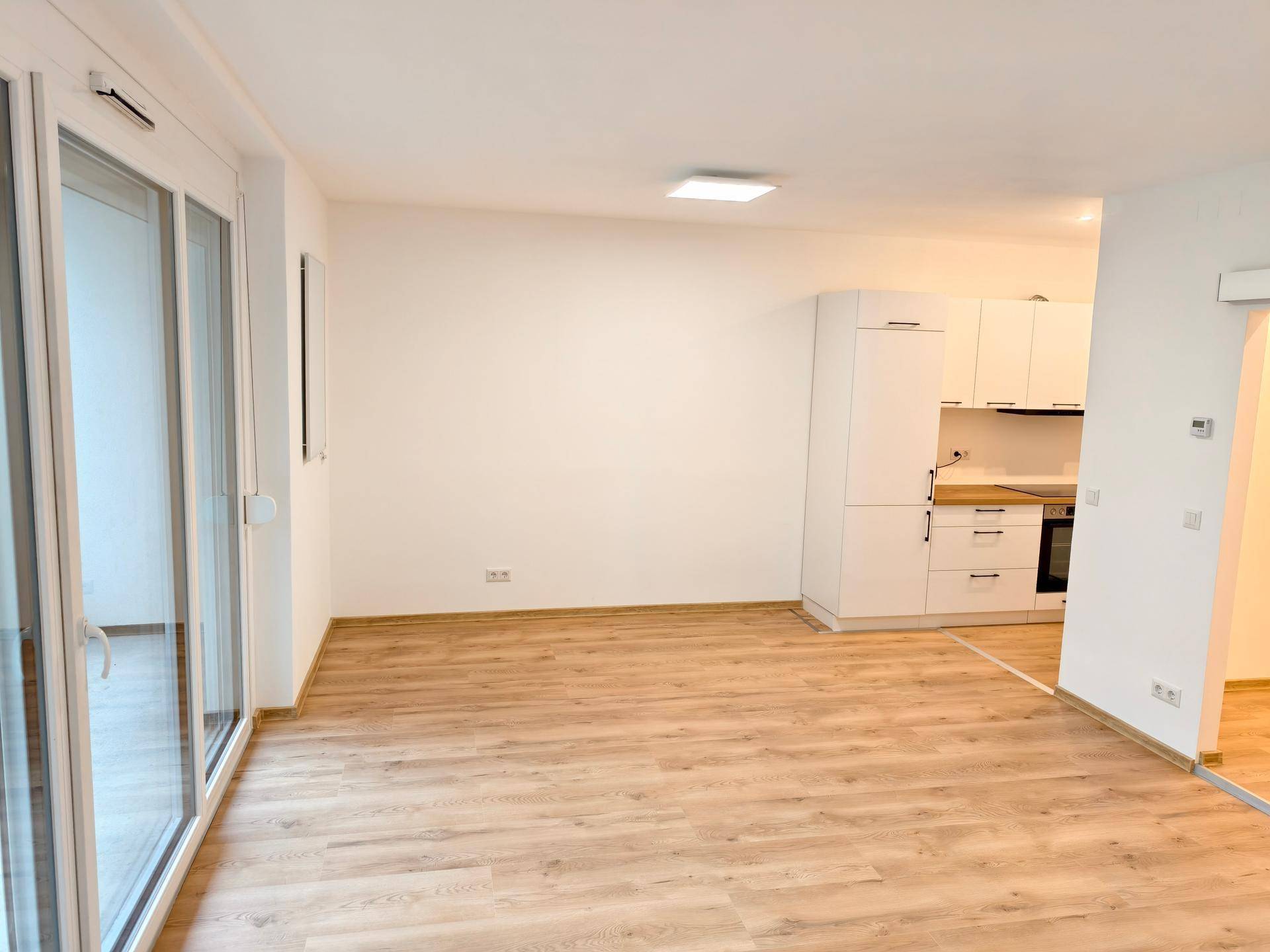 Heller Wohnbereich mit Laminatboden, offener Küche im Hintergrund und großem Fenster mit Zugang nach draußen.