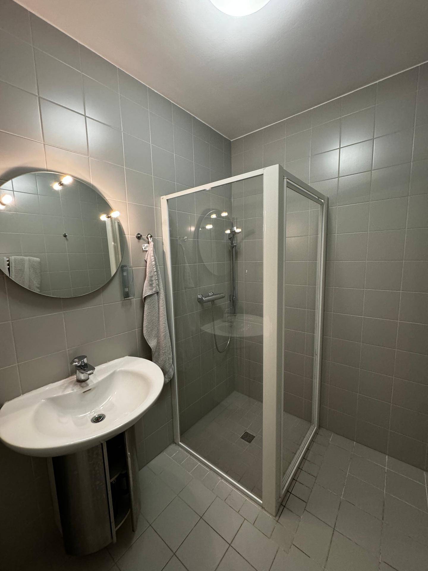 Modernes Badezimmer mit grauen Fliesen, Duschkabine und rundem Spiegel.