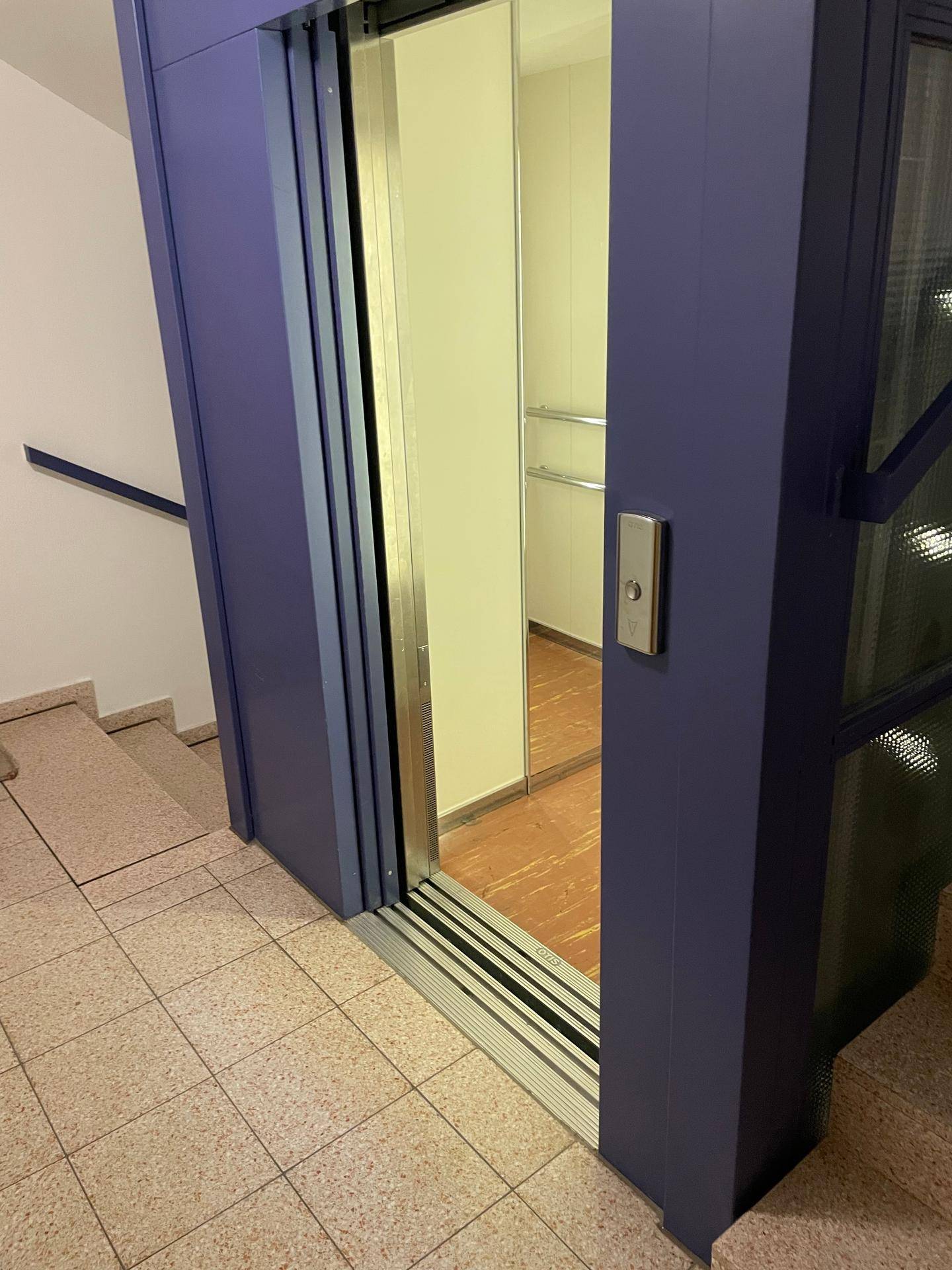Moderner Aufzug mit blauer Verkleidung und Treppenhaus mit Fliesenboden.