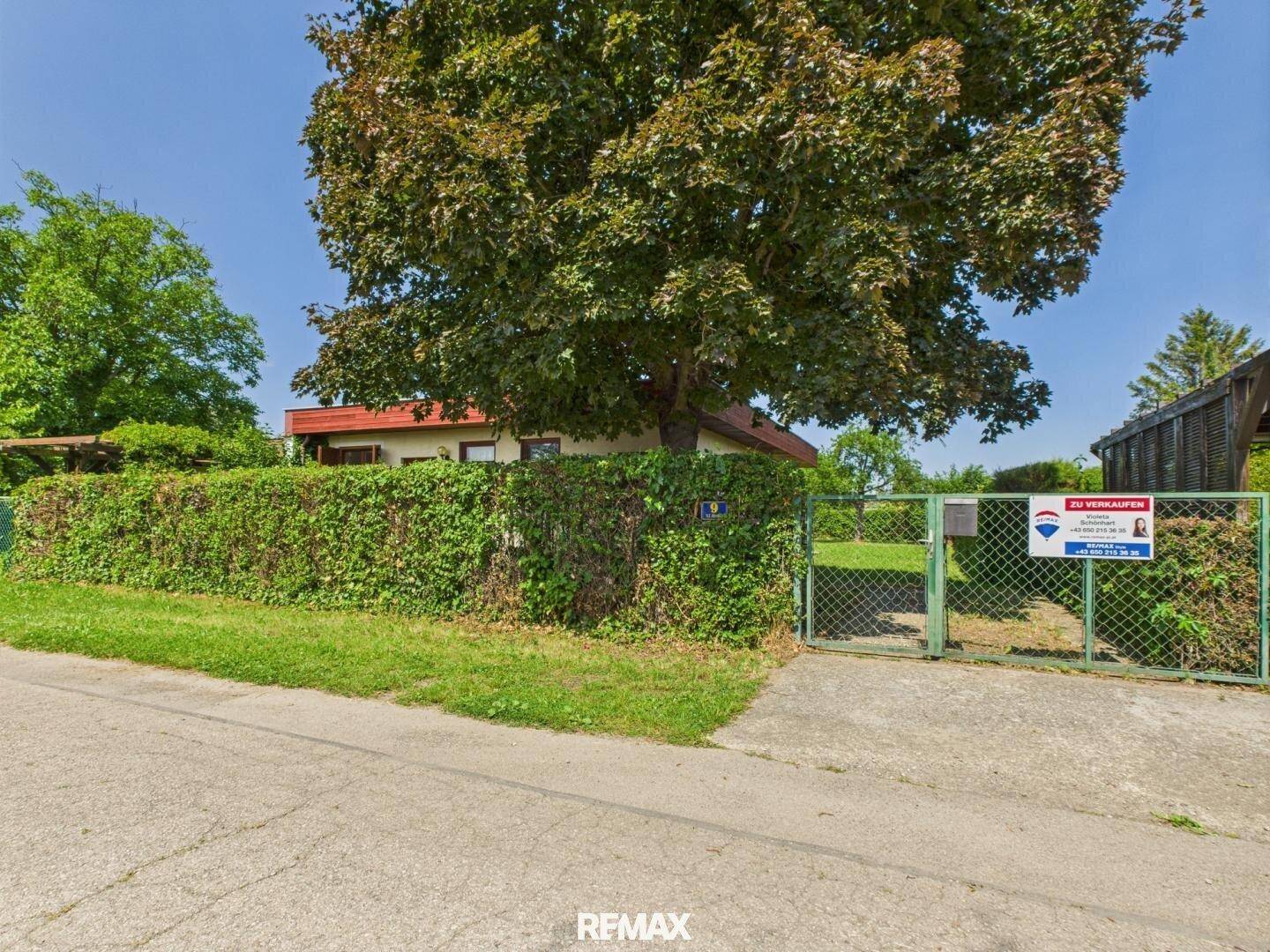Eingangsbereich des Grundstücks von der Straße aus mit Tor, Hecke und einem Verkaufsschild.