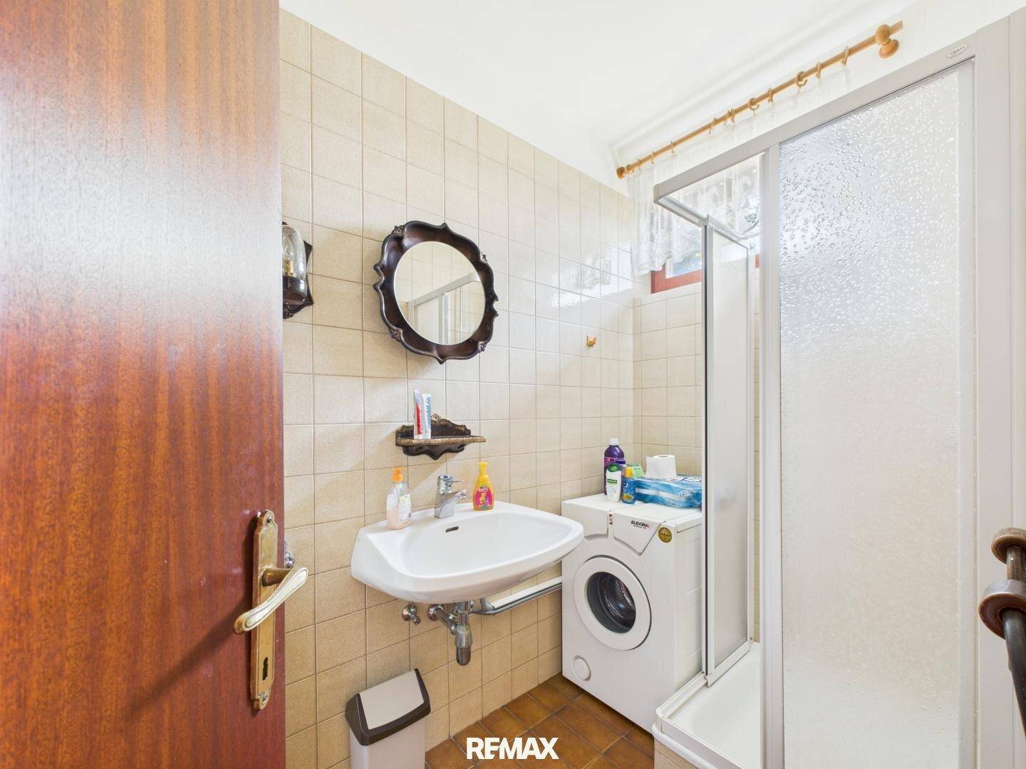 Badezimmer mit hellen Fliesen, Waschbecken, Waschmaschine und einer Duschkabine.