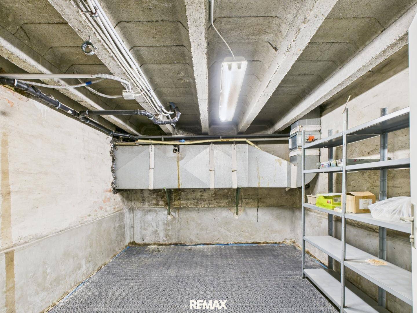Geräumiger Kellerraum mit Betonwänden, Regalen und freiliegenden Rohren an der Decke.
