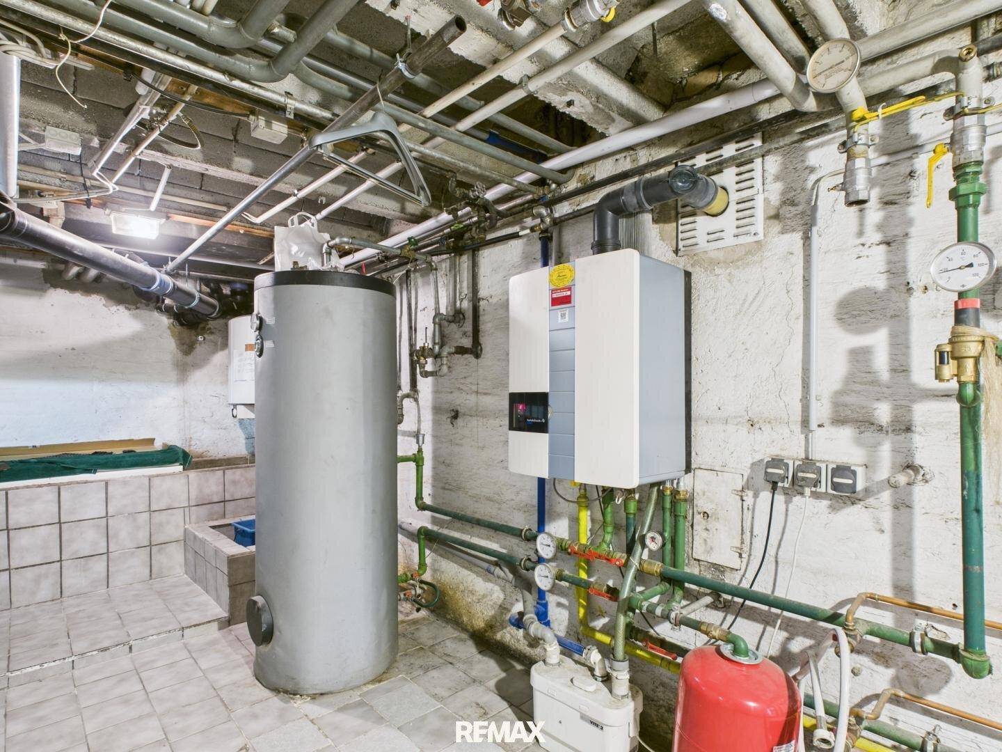 Heizungsraum im Keller mit Warmwasserspeicher, Heizkessel und zahlreichen Rohrleitungen.