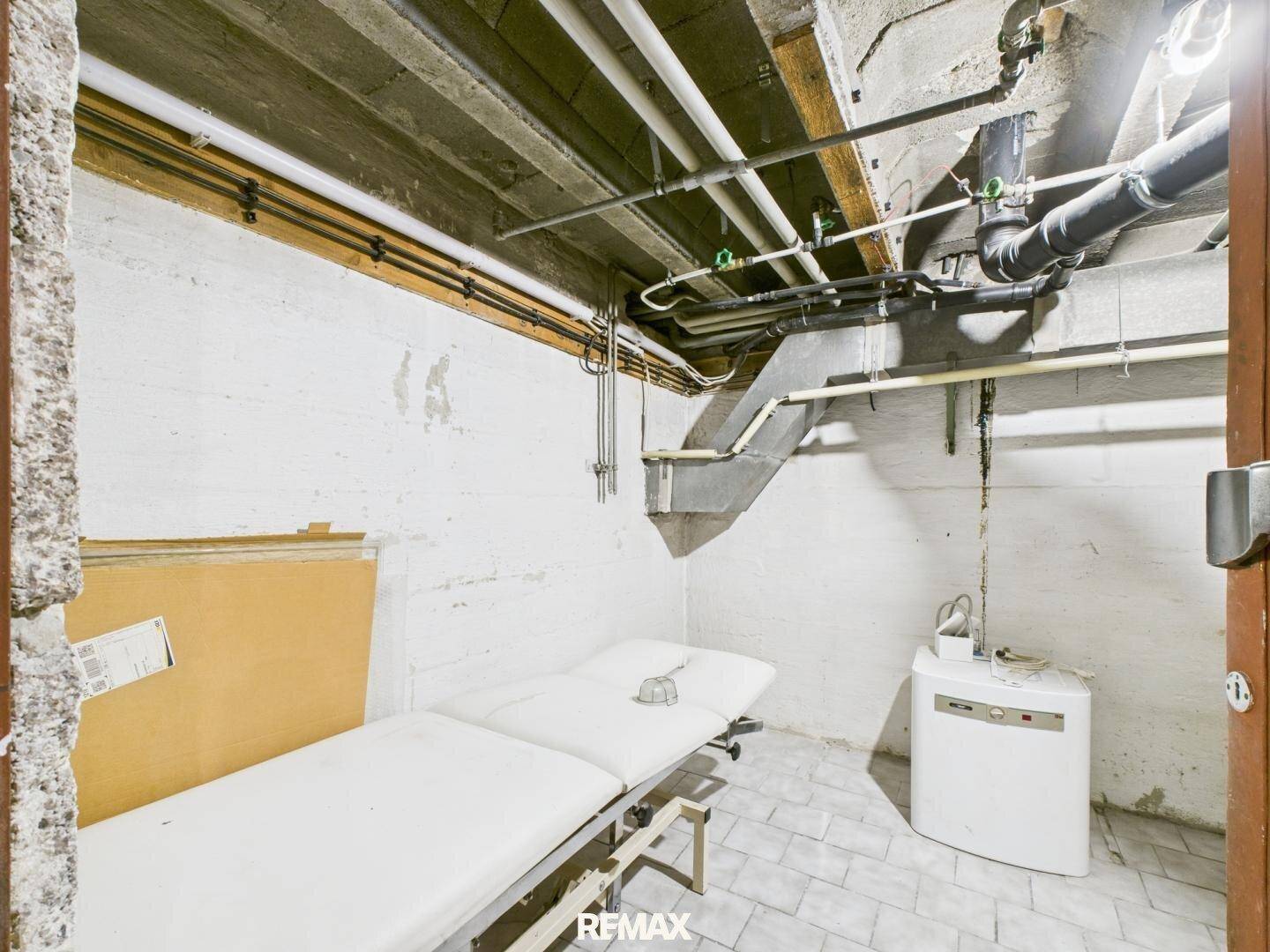 Kellerraum mit Fliesenboden, einer Liege und freiliegenden Rohren an der Decke.