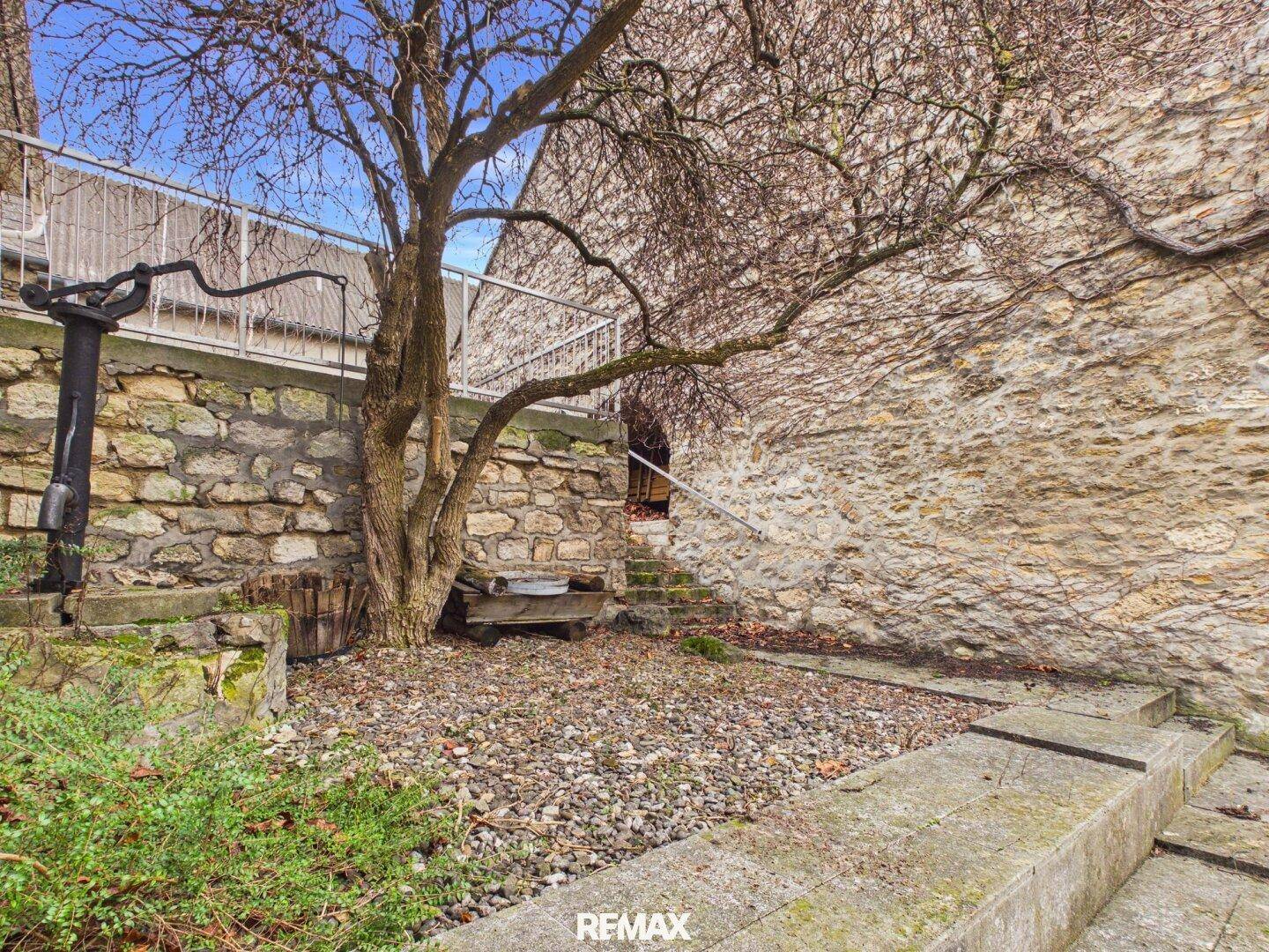 Historischer Gartenbereich mit alten Steinmauern, einem Baum und Treppen, die zu einem Eingang führen.