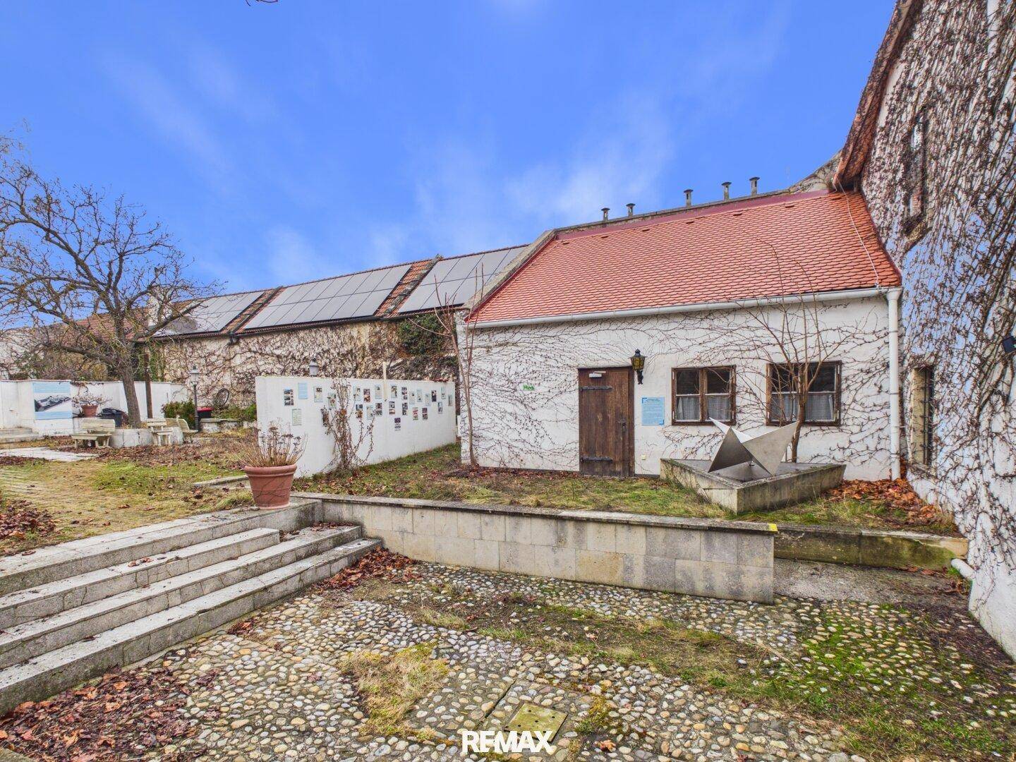 Innenhof mit einem kleinen Gebäude, Kieselsteinweg und einer alten Mauer mit Solarpaneelen im Hintergrund.